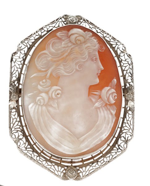 Cameo Brooch in 14 Karat : A 14k white gold shell cameo brooch depicting a woman facing left. The cameo measures 44mm x 33mm with floral accents. The surrounding mounting features filigree work with floral accents. 8.2 dwt.