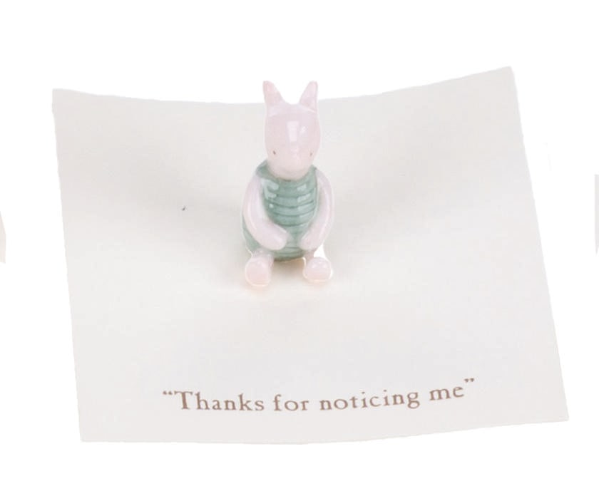 A Mini "Winnie the Pooh" Piglet Figurine.: (Michel and Co., 1990s) A mini ceramic figurine of Piglet, inspired by the original illustrations for the Winnie the Pooh books by E. H. Shepard. The figurine is attached to a card with a quote that
