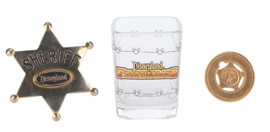 A Trio of Frontierland Souvenirs.: (Disneyland, 1980s-90s) Three Frontierland souvenirs, including a shot glass, typer token, and a sheriff's badge. The sheriff badge is very similar to the Park badges, where Guests could have their