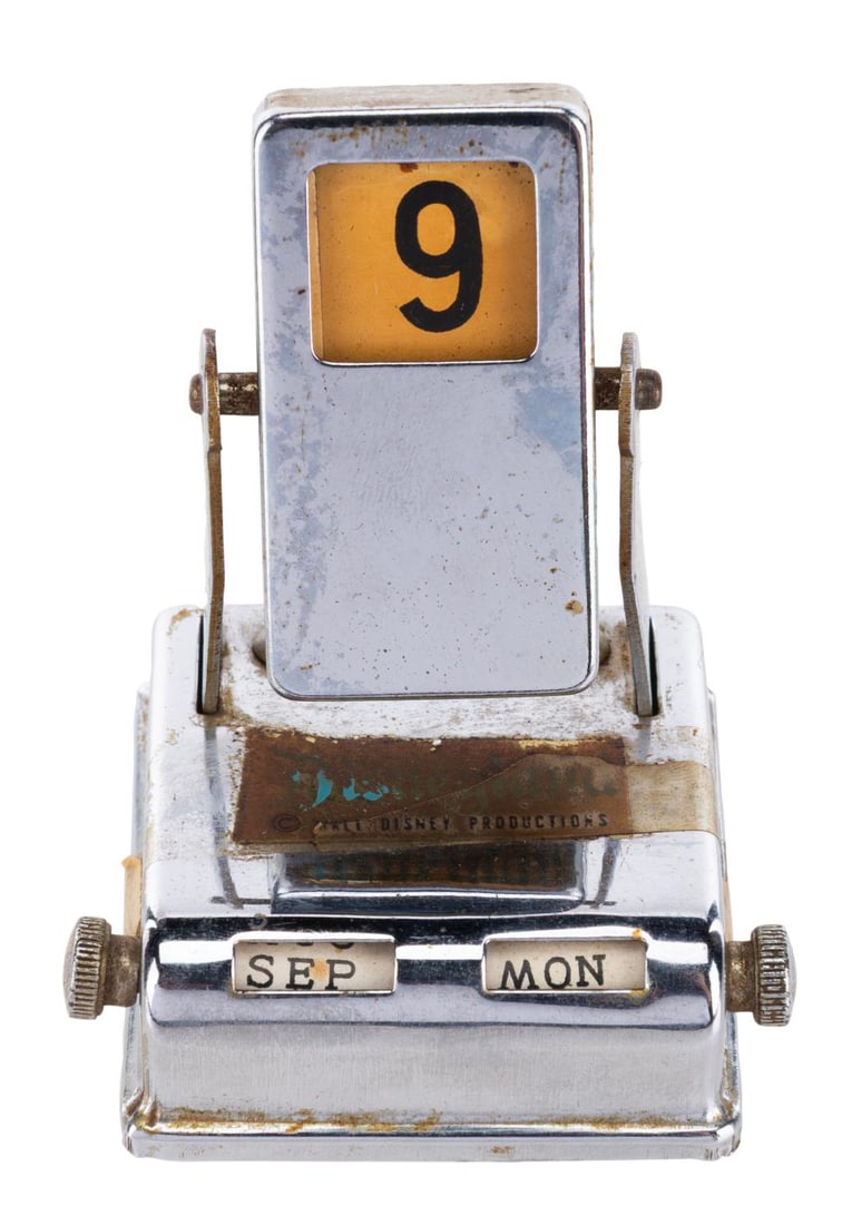 A Souvenir Perpetual Calendar with Disneyland Decal.: (Disneyland, 1960s) A silver-tone perpetual calendar with a Disneyland decal. The months and days can be changed by the scroll wheels on the base, and the date changes when the top piece is rotated.