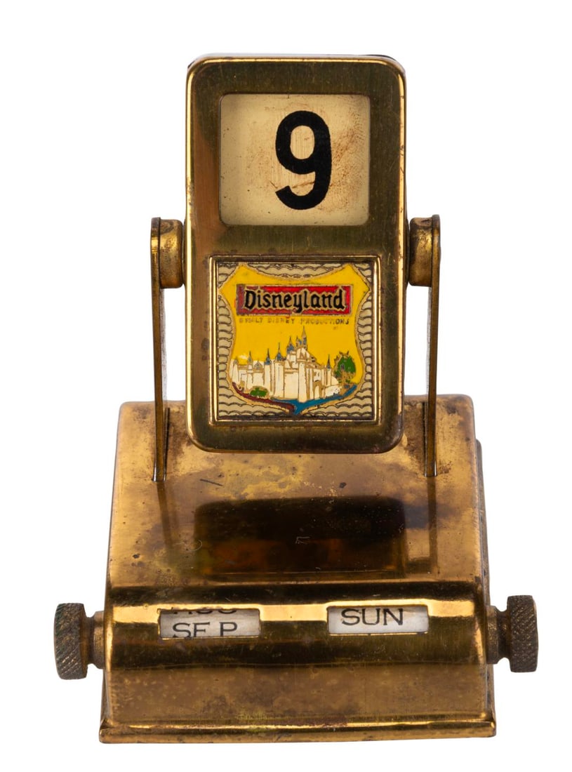 A Gold-Tone Disneyland Perpetual Calendar.: (Disneyland, 1950s-60s) A Disneyland gold-tone perpetual calendar. The months and days can be changed by the scroll wheels on the base, and the date changes when the top piece is rotated. Measuring 2.