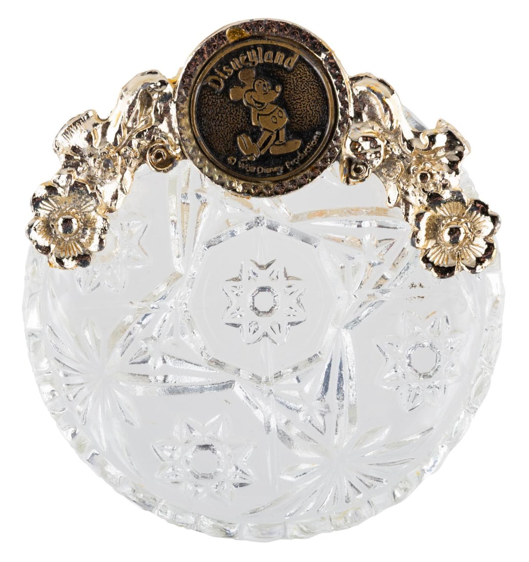 A Disneyland Souvenir Trinket Dish.: (Disneyland, 1970s) A souvenir trinket dish featuring a Disneyland Mickey Mouse emblem. Measuring 4" in diameter, the glass dish with plastic emblem is in very good condition .