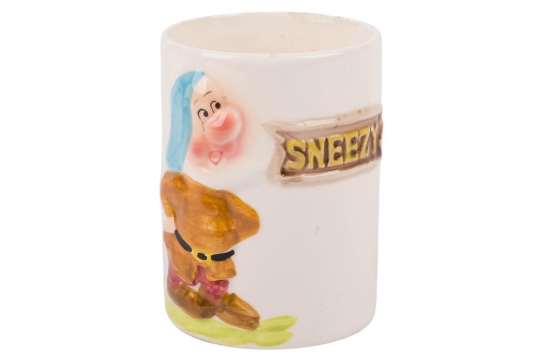 A "Snow White" Sneezy Ceramic Cup.: (Enesco, 1960s) A ceramic cup with a 3D relief of Sneezy from "Snow White and the Seven Dwarfs." Measuring 2.75" in diameter and 3.75" tall, the cup is in fair condition with a chip on the rim and cra