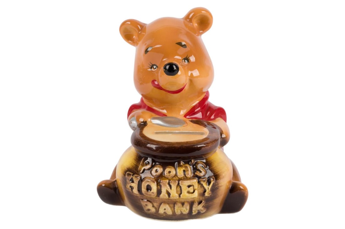 A Ceramic Winnie the Pooh Savings Bank.: (Enesco, 1960s) A ceramic figural savings bank of Winnie the Pooh with a pot of honey, manufactured by Enesco. Measuring 4" x 4" and 4.75" tall, the bank is in very good condition with light wear