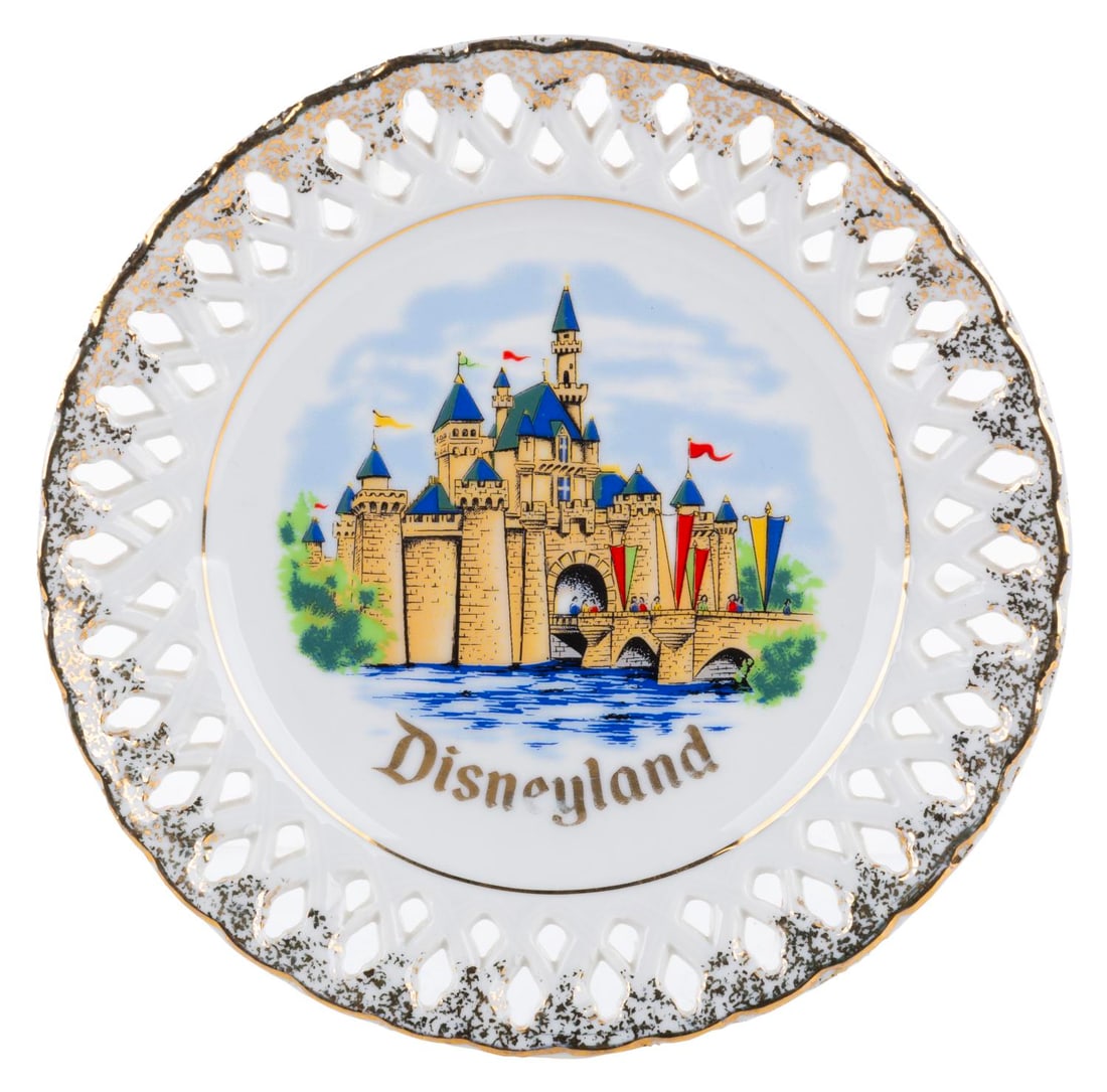 A Decorative Lace Sleeping Beauty Castle Plate.: (Disneyland, 1970s) A souvenir decorative lace plate featuring Sleeping Beauty Castle and delicate gold detailing. Measuring 6.25" in diameter, the plate is in very good condition with light fading