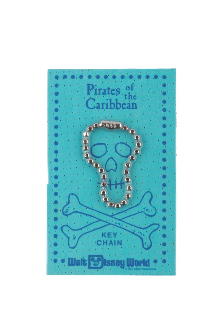 An Early Pirates of the Caribbean Skull Keychain.: (Walt Disney World, 1970s) A souvenir Pirates of the Caribbean keychain, originally distributed in 1968 with the intention of being used for the Park's souvenir doubloons. The packaging presents the s