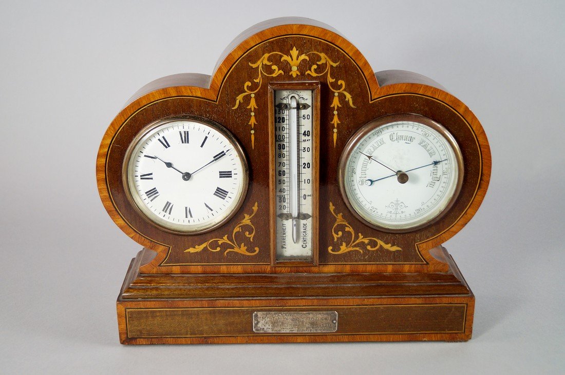 An Edwardian mahogany and inlaid clock/barometer, the: An Edwardian mahogany and inlaid clock/barometer, the case of clover leaf outline, decorated with line inlaid and tendril decoration, with a central thermometer flanked by a clock with white enamel di