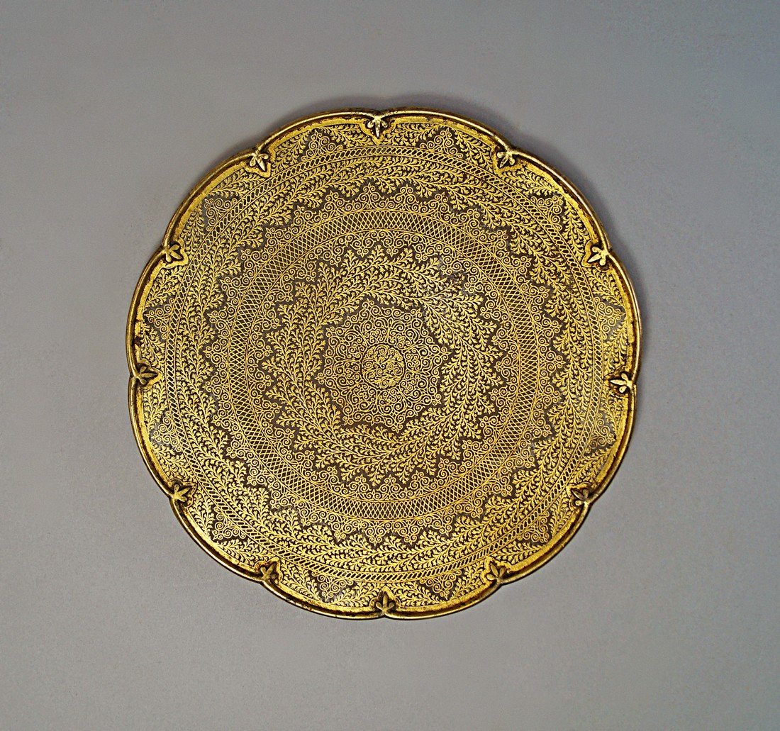 An Indian circular damascened plate, 19th century, with: An Indian circular damascened plate, 19th century, with shaped petal outline rim, inlaid to the main body with concentric bands of trailing tendrilous plants and geometric designs, the base with etche