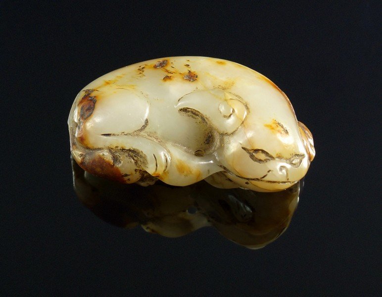 A Chinese carved pale and russet green recumbent lion,: A Chinese carved pale and russet green recumbent lion, Late Ming dynasty, reclining and with curved body and carved detail all around, with stylised features and typical surface deposits, 3.5cm wide
