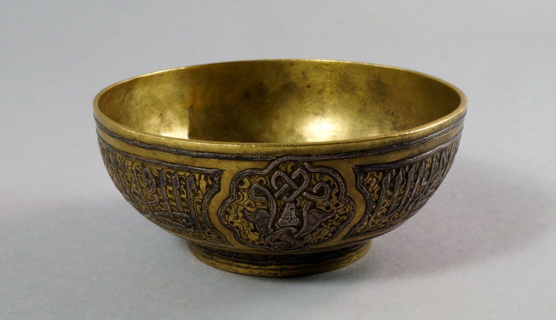 A Mamluk style calligraphic white metal inlaid brass: A Mamluk style calligraphic white metal inlaid brass footed bowl, 18th/19th century, of tapering and circular form, decorated with panels of script flanking lobed panels of foliage, 10.5cm diam