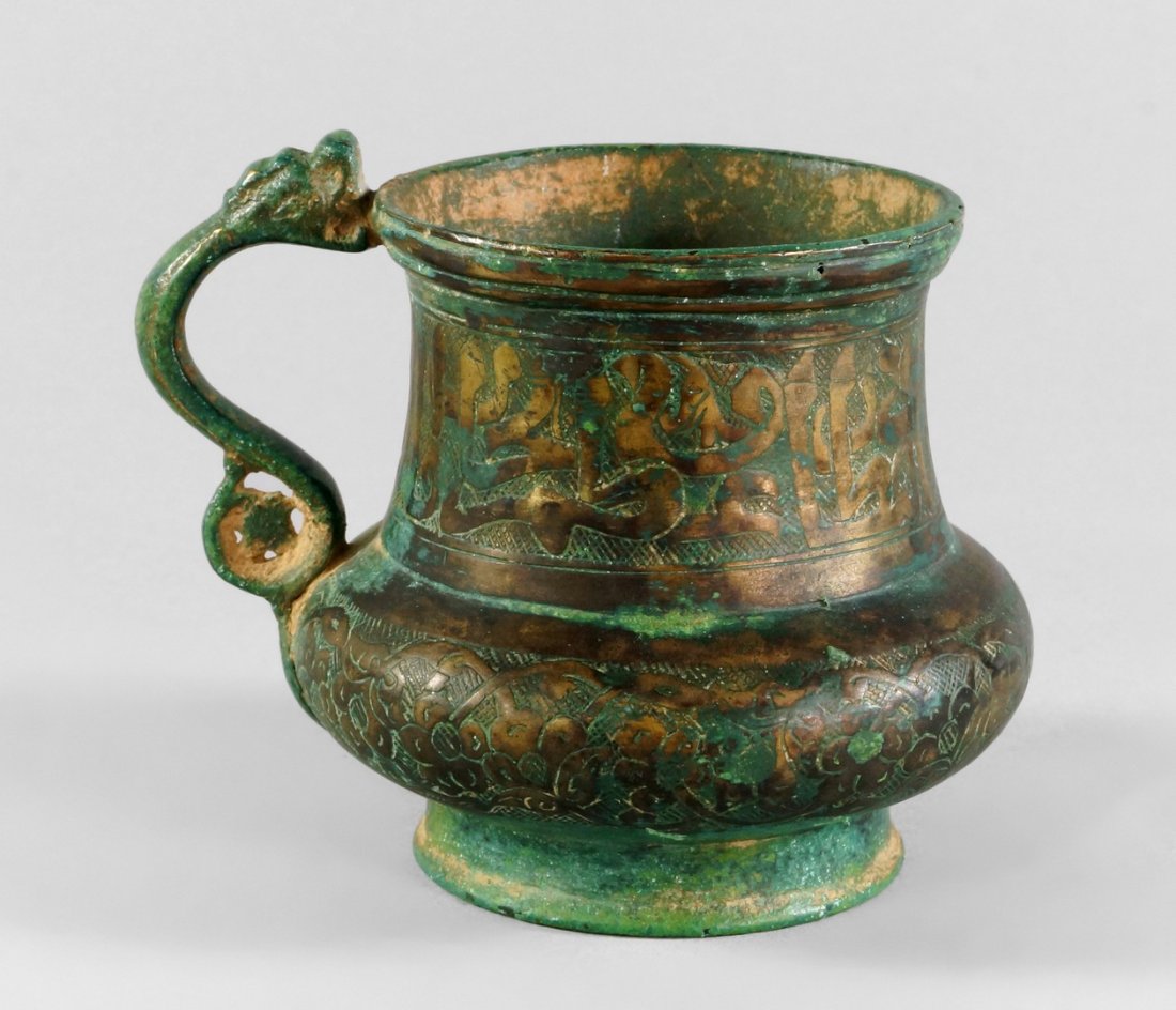 A bronze cup, Timurid, 14th century, of squat form with: A bronze cup, Timurid, 14th century, of squat form with scrolling handle terminating in a dragon head, the neck worked with bands of calligraphy, the bulbous belly with a band of foliage and flower he