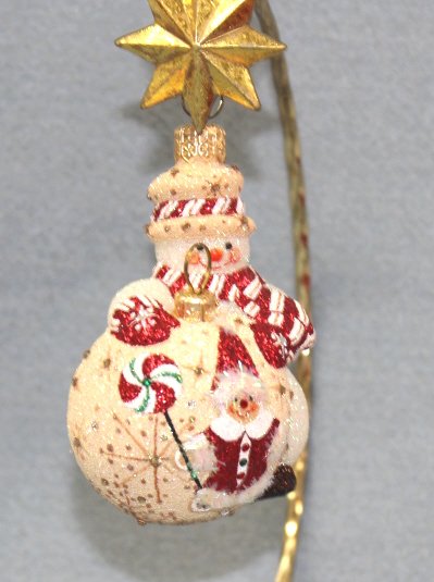 Patricia Breen "Under Construction-Gingerbread" Or: Patricia Breen "Under Construction Gingerbread" Christmas ornament. White snowman with a gingerbread hat trimmed with a red, white, gold candy cane stripe and matching scarf. He is rolling out a ginge