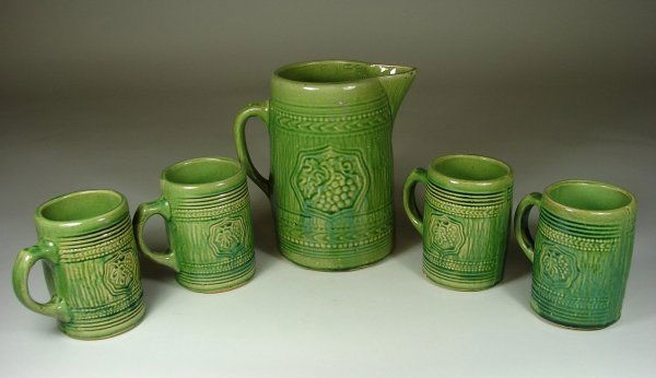 Green Salt Glazed Pitcher & 4 Mugs: Grape Cluster in Shield pattern, pages 47 & 49 in Salt Glazed Stoneware by Taylor & Lawrance. Light glaze crazing, one small shallow flake on one handle. Pitcher 8 3/4" h x 5 3/4" dia.