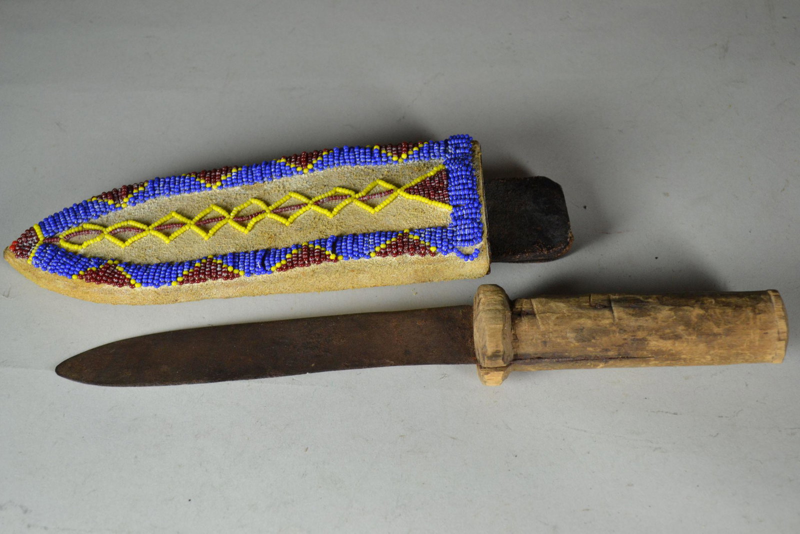 Crude Handmade Knife: with thin steal blade in beaded scabbard. Wood grip is hand carved and shows heavy wear. Blade is rusted. Leather scabbard has been covered in beaded rawhide and sewn with sinew. Moderate wear to scab