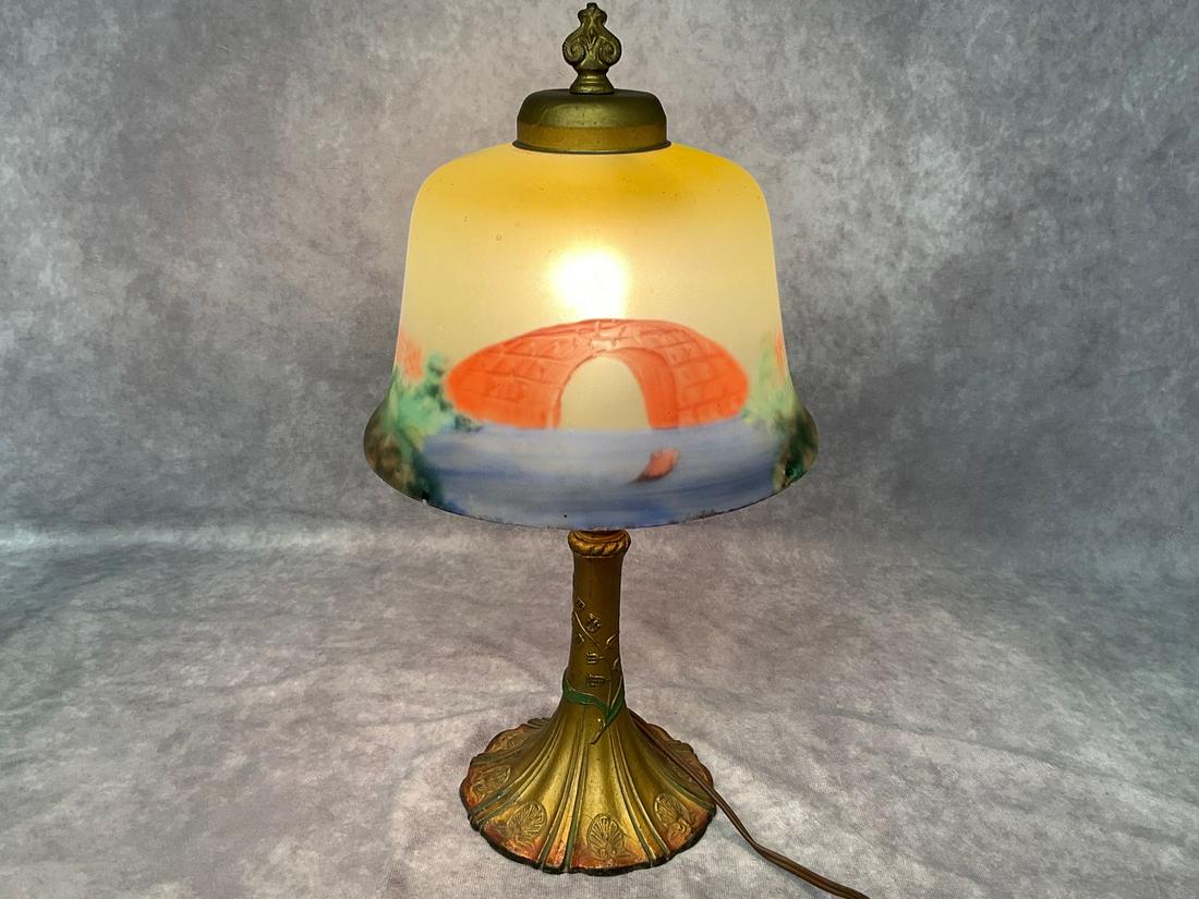 Reverse Painted Boudoir Lamp: Polychrome cast iron base. Shade with bridge and woodland scenery. Edges of glass shade is chipped in spots. Powers on. 15 inches tall.