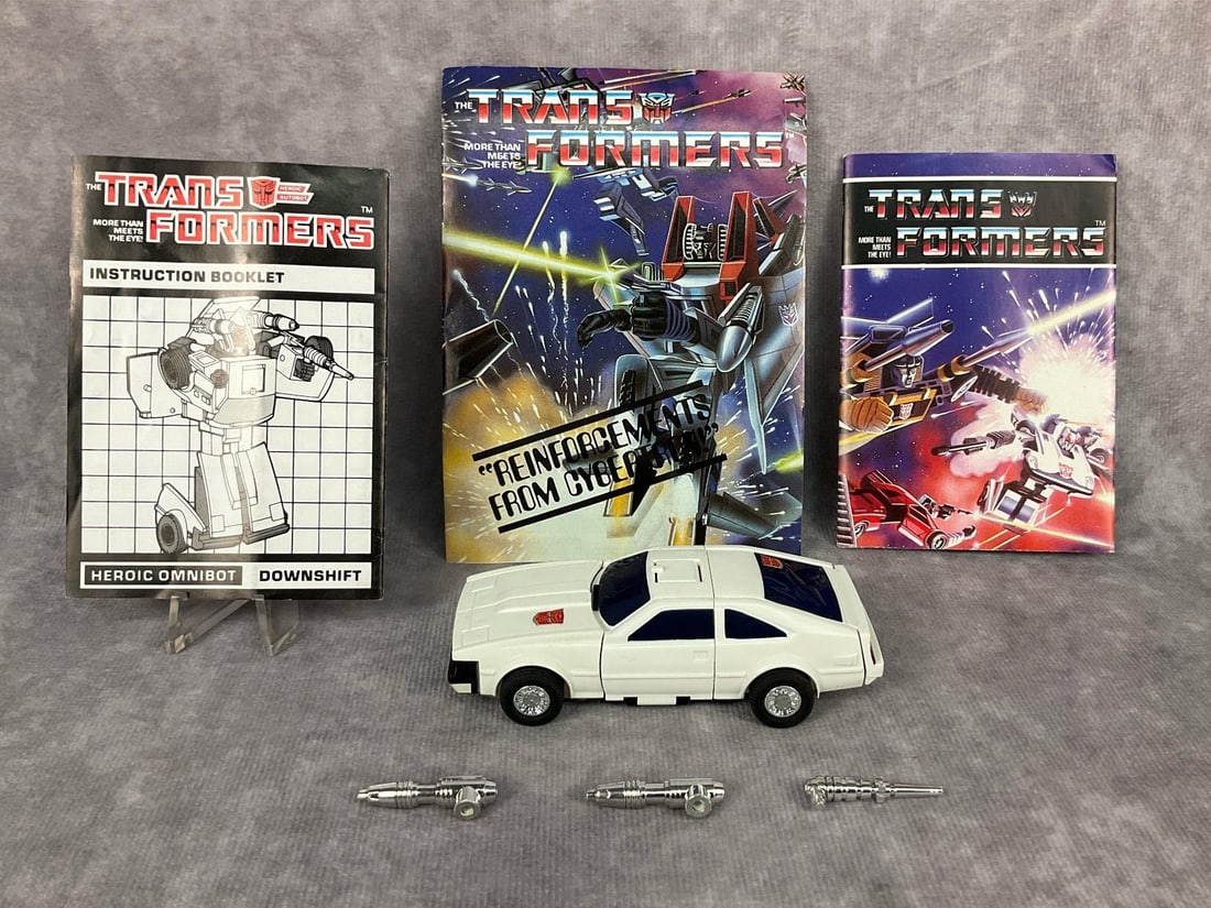 1985 Downshift Transformer: Car is marked Tamara Japan, booklets are by Hasbro. Includes instruction booklet, catalog, mini poster, and weapons. Missing tech spec card and decoder. Good condition. Transformer measures 4 inches l