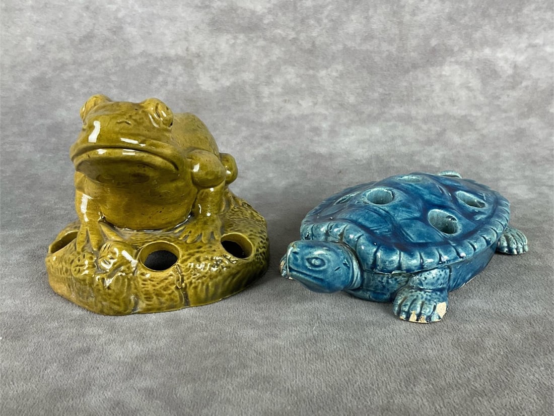 Majolica Flower Frogs: One green frog flower frog with six holes, one blue turtle flower frog with seven holes. The turtle has chips on the feet, and the frog is in good condition. Turtle measures 5.5 inches wide x 1 inch t