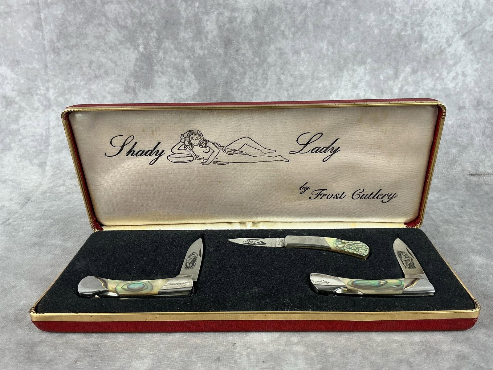 Shady Lady Set by Frost Cutlery: Group of three foldable pocket knives with nude women on the blades. Good condition. The largest knife measures 3 inches wide, closed. Case measures 10 inches wide x 4 x 1 inch tall.