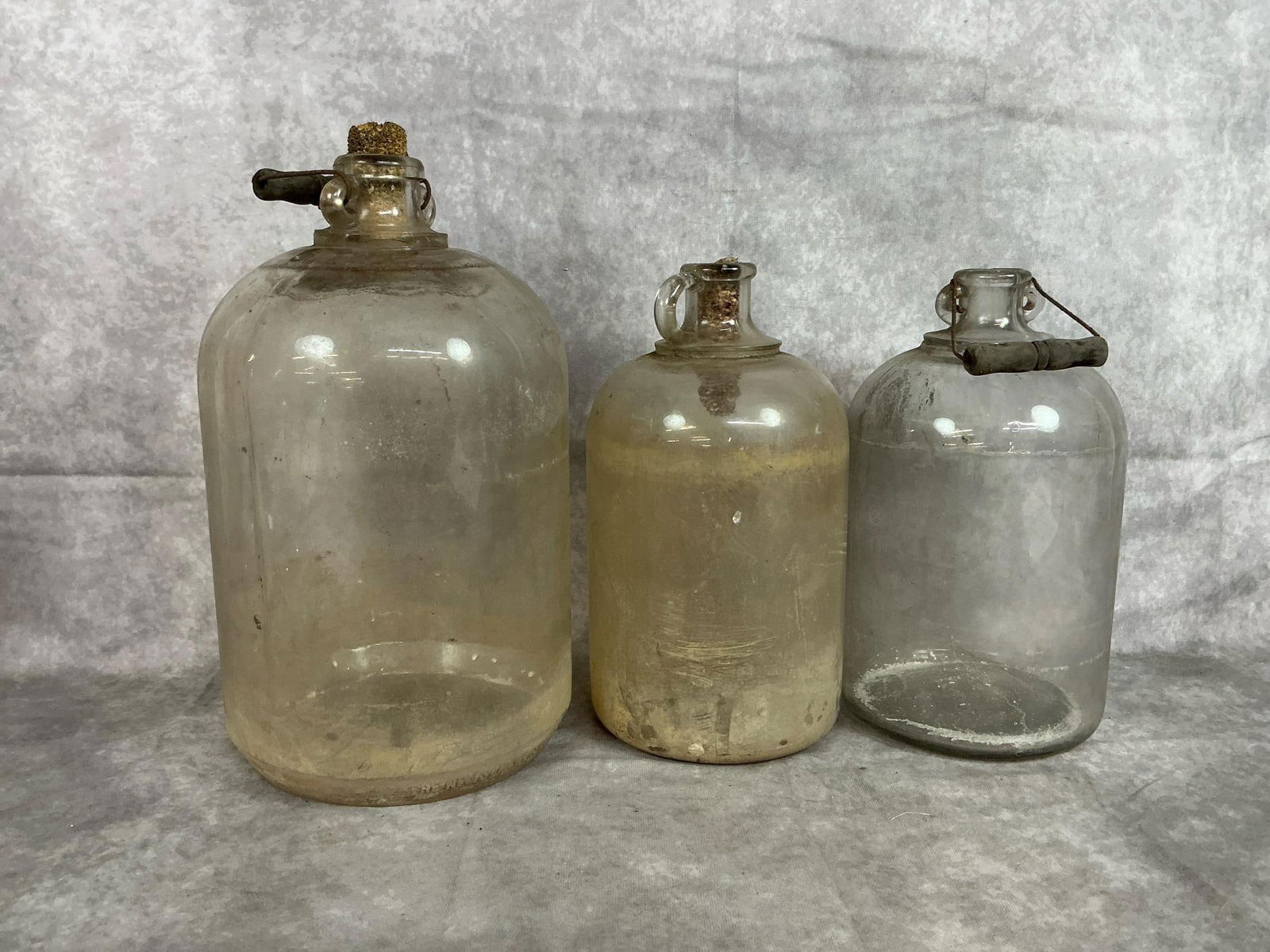 Moonshine Jugs: Three early 1900s jugs. One 2-gallon glass jug with handle from The Wivison Weiskop Company. Two 1-gallon glass jugs with finger handle and wire bale wooden handle. Handles are loose and jugs are