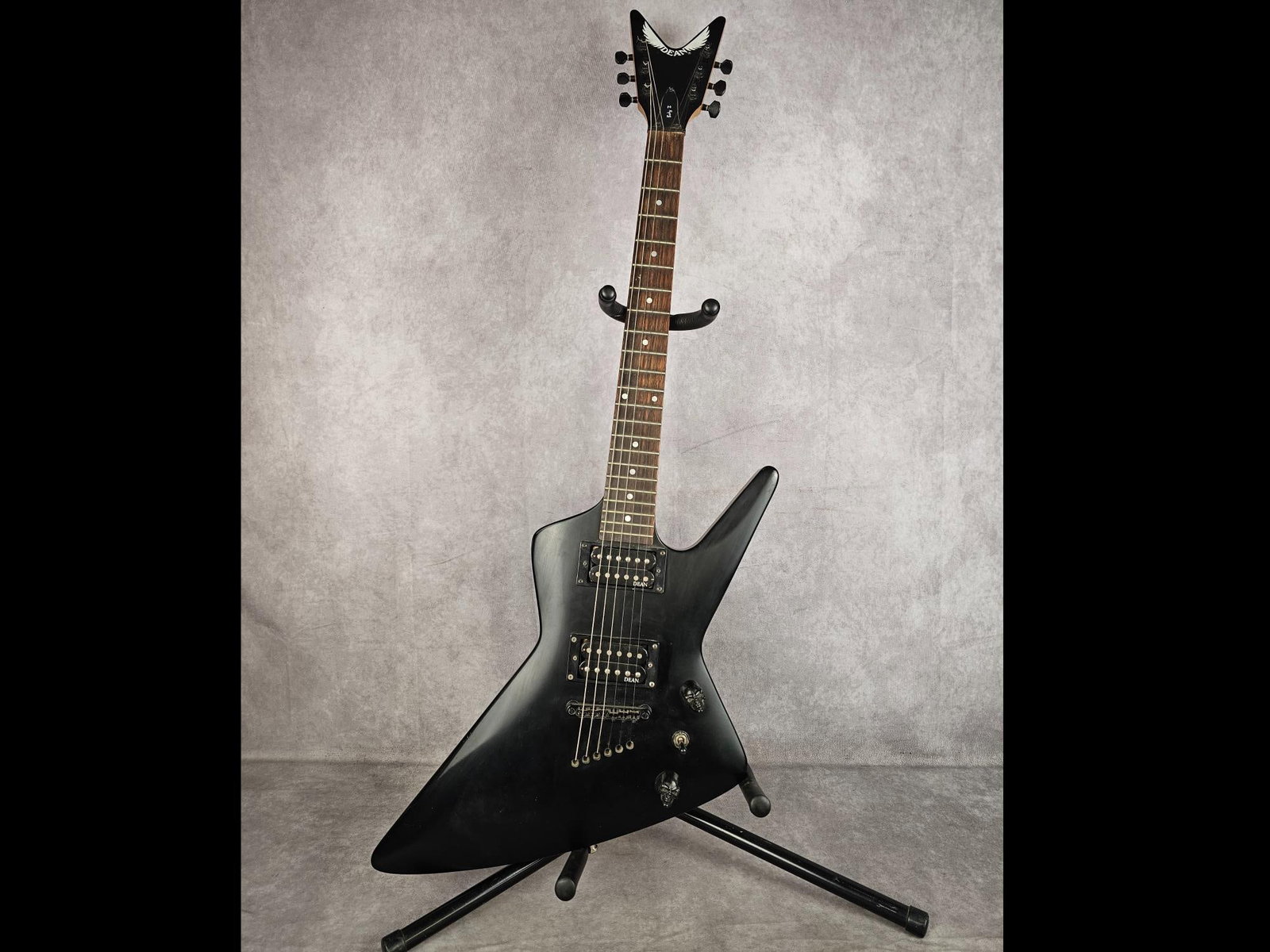 Dean Baby Z Electric Guitar: Matte black angular, explorer style body, six string electric guitar. Features dual humbucker pickups, skull volume nobs, solid wood neck, V shape headstock. Serial number 0550344, made in China. Brid