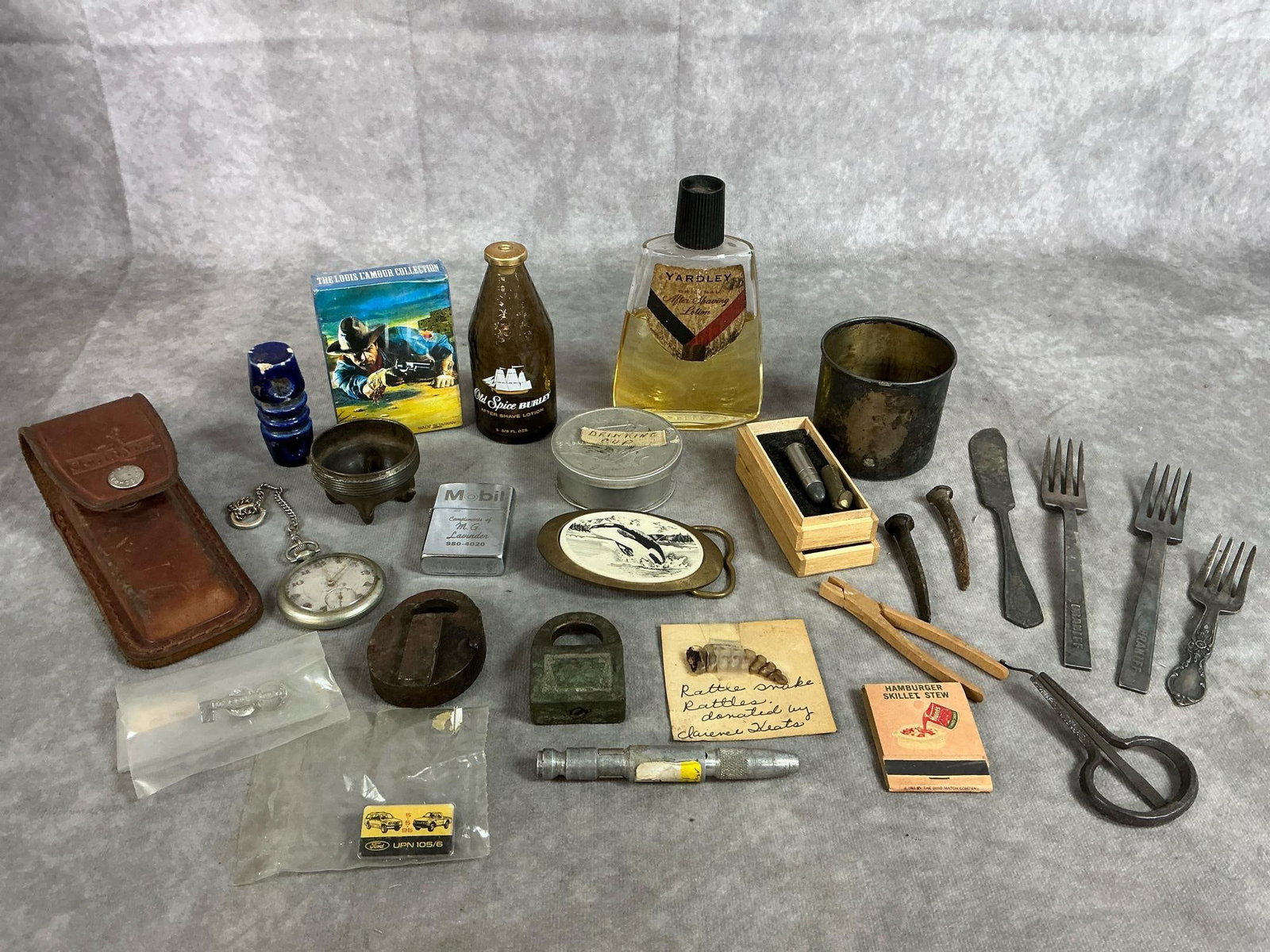 Vintage Collection of Men's Finds: Twenty nine early items. Four pieces of silver plated flatware, bullets, rattlesnake rattler, two locks, and vintage after shave. Includes Louis L'Amour playing cards, pocket watch, and two Ford tac