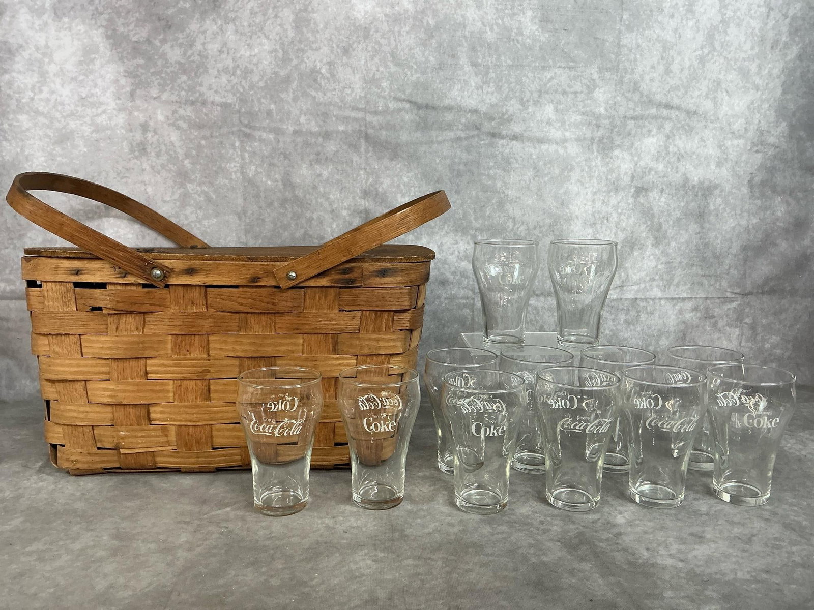 Vintage Picnic Basket & Coca-Cola Glasses: Picnic basket and a dozen glasses from the mid 1900s. Woven wood splint basket with hinged lid and handles. Twelve 5” high Coca-cola glasses. Basket shows signs of wear and broken splints in