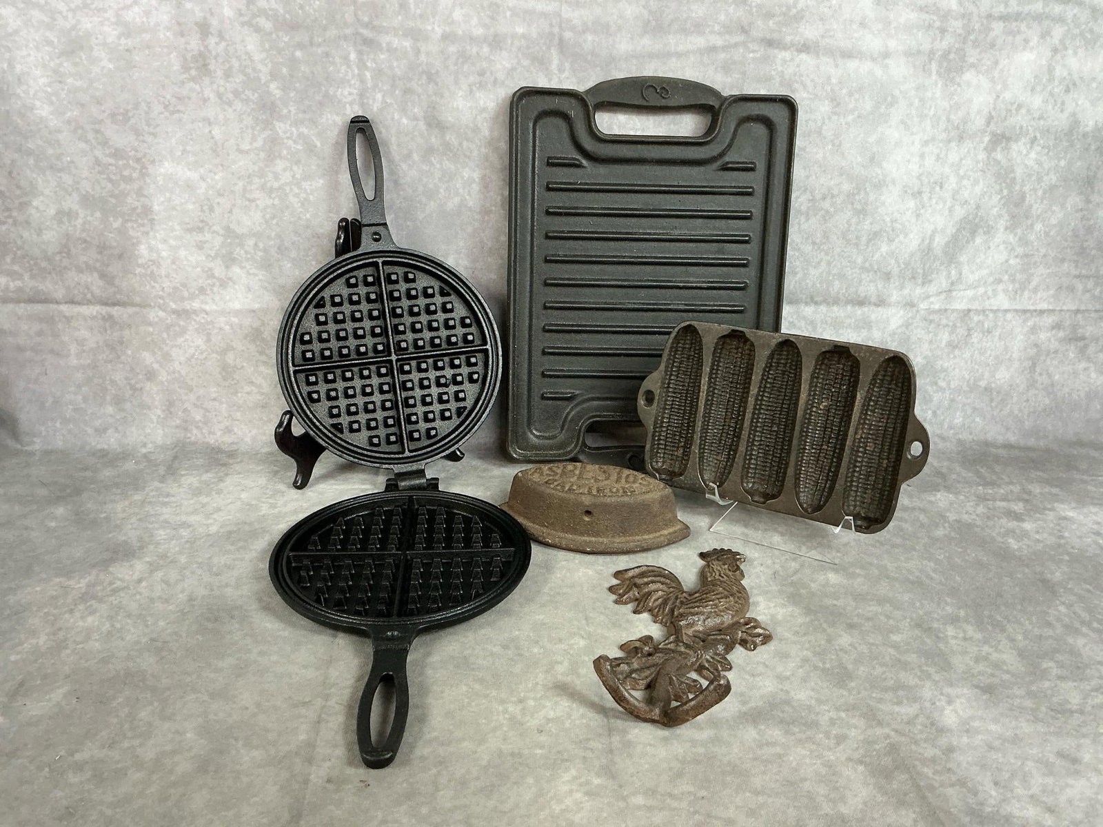 Cast Iron Collectibles: Five early to late 1900s pieces. Sugarcreek Supply waffle griddle, rectangular grill, and cornbread pan. Includes Asbestos sad iron and rooster wall hook. Some signs of rust, otherwise in good conditi