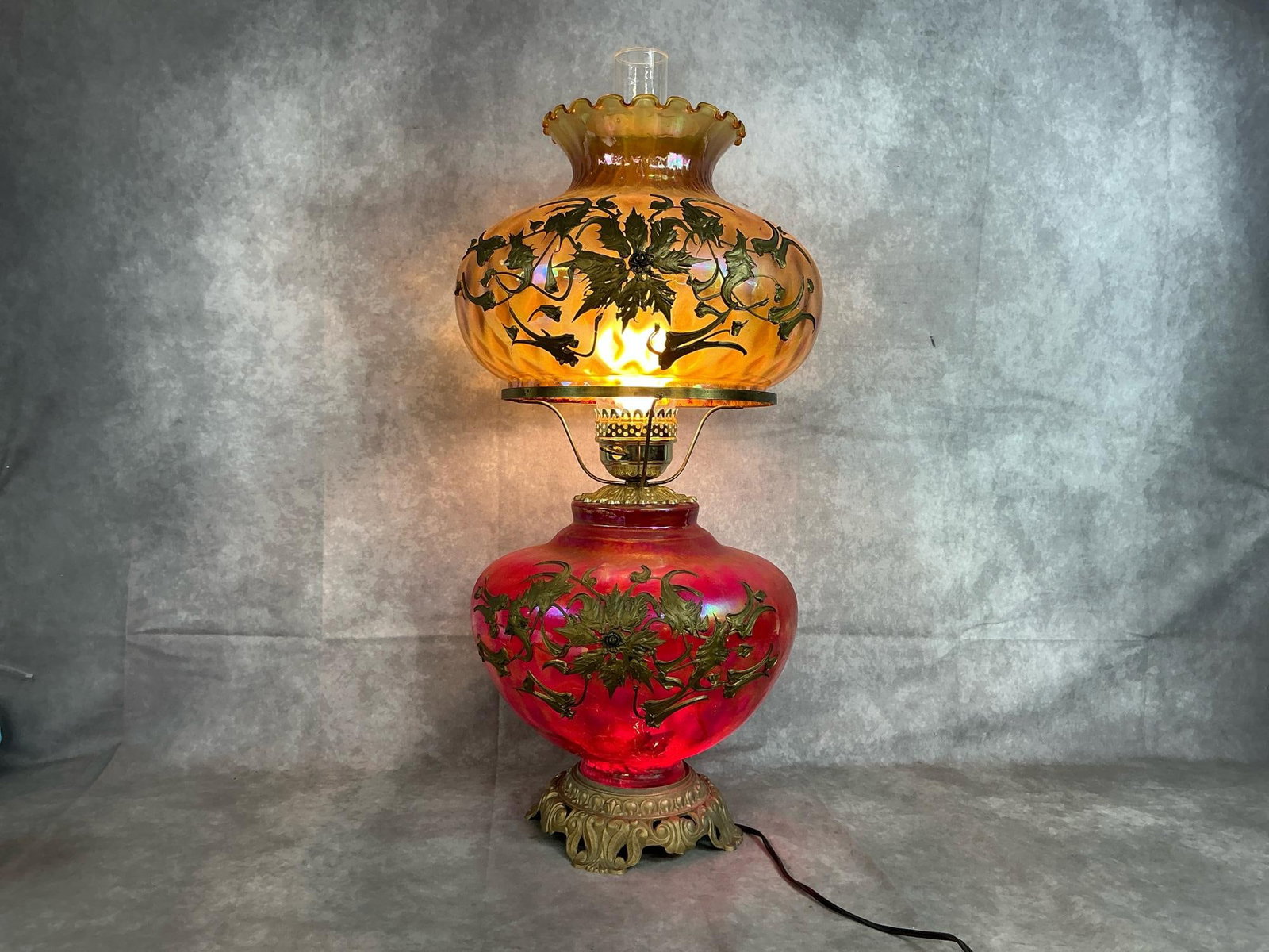 Victorian Parlor Lamp: Antique two bulb parlor lamp. Cranberry glass base with textured gold glass top globe. Both have applied brass floral motifs and a clear glass chimney. Some crazing and discoloration. Lamp measures