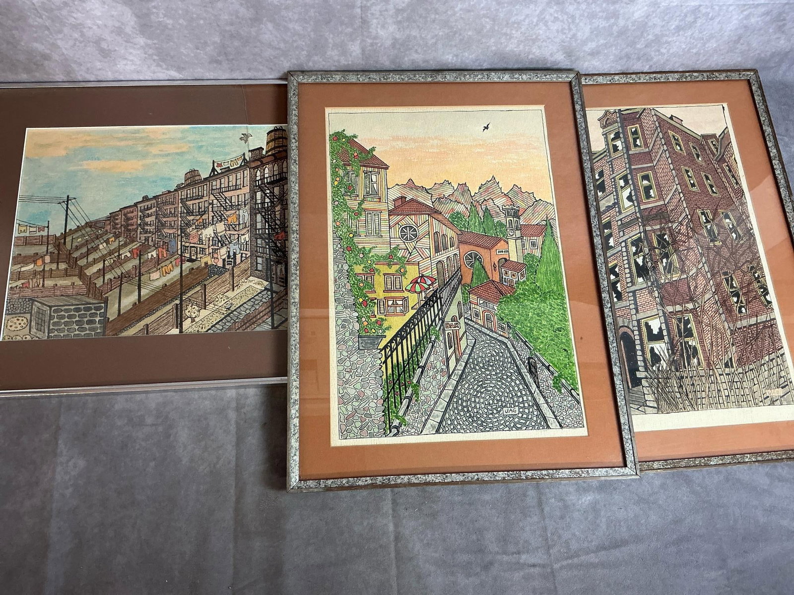Watercolor Community Scenes by JAG: Three framed prints by JAG. Variety of detailed watercolor style "buildings". Tan matting. One glass is broken. Picture measures 15 inches wide x .5 x 21 inches tall.