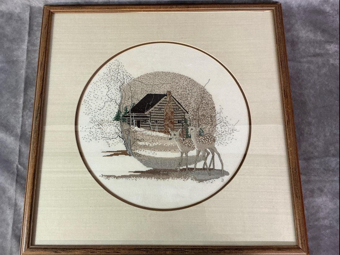 P Buckley Moss designed "Mountain Lodge" Cross-stitched: 1991 cross-stitched framed and matted cabin and deer picture. Inspired by P Buckley Moss, with tans, browns, black, and green colors. In good condition. Measures 22 inches wide x 1 x 22 inches tall.