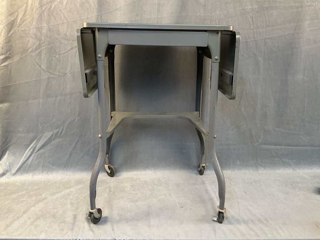 Typewriter Table: Mid-20th century vintage metal office typewriter table with castors and drawer. Includes two period 1940's typing books. Good condition. Measures 19.5 inches wide x 14.5 x 26 inches tall.
