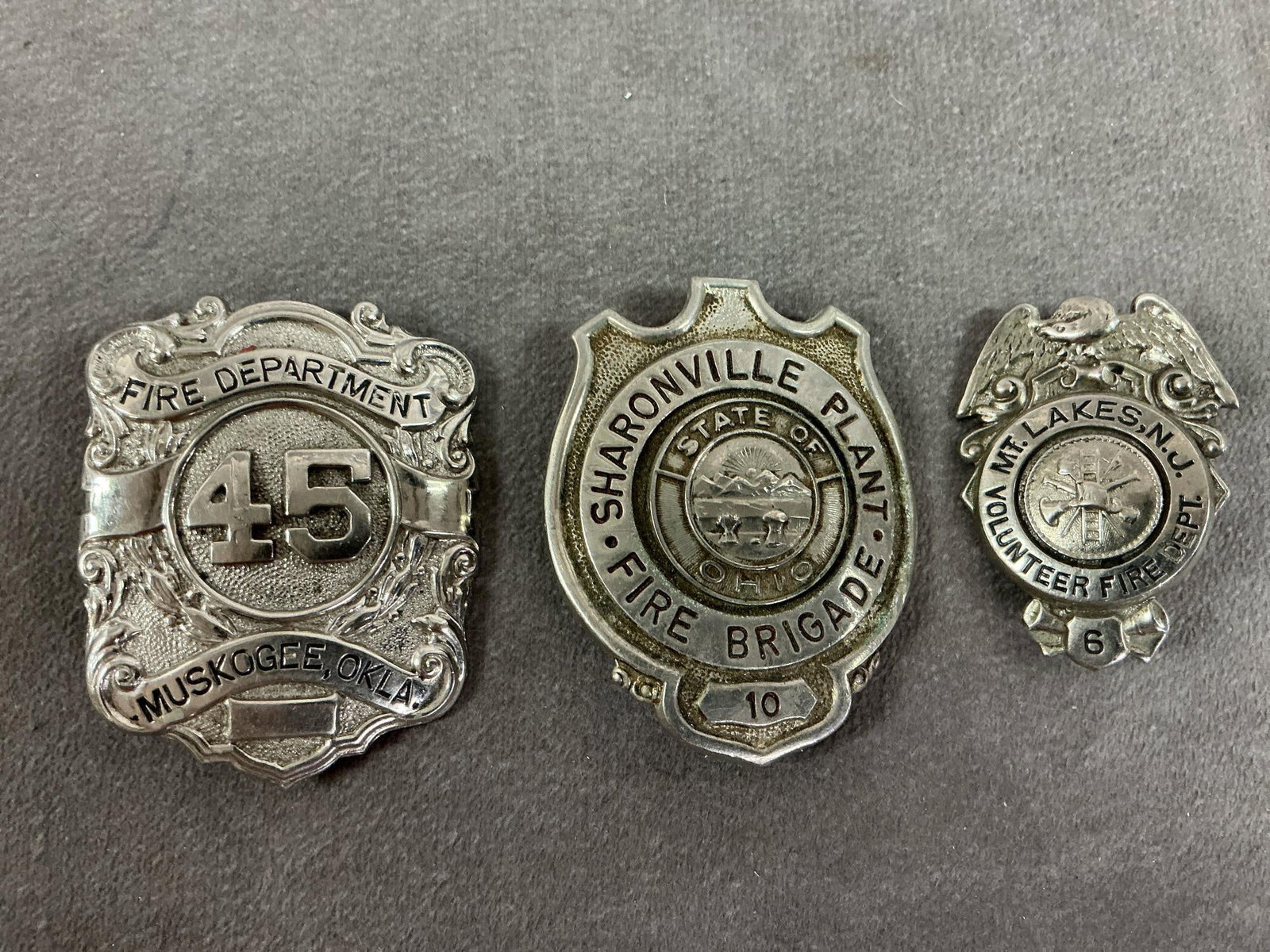 Fire Department Badges: Three vintage badges. Sharonville, Ohio Fire Brigade, Muskogee, OK, and Mt Lakes, NJ fire departments. In good condition. Largest measures 1.5 inches wide x 2.25 inches tall.