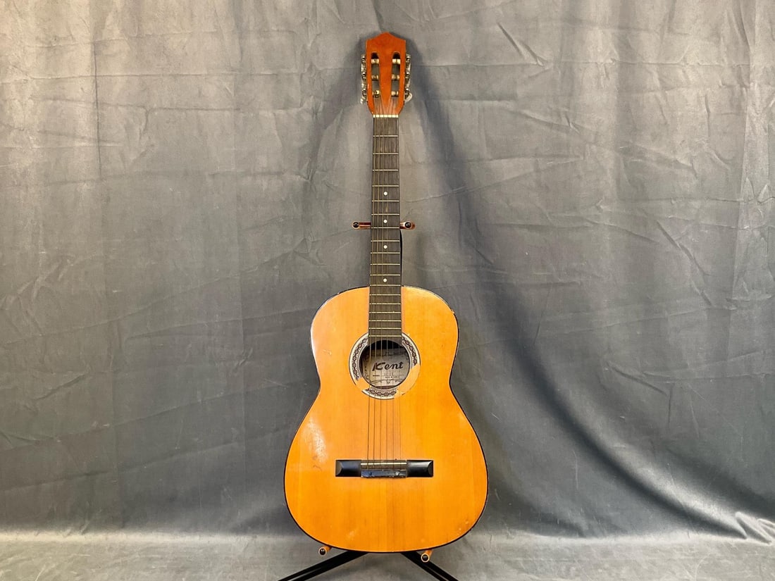 Kent Acoustic Guitar: Late twentieth century vintage Kent model KC 130 acoustic guitar with stand. Scrapes, gouges, and missing decorative decal around sound whole. Measures 14 inches wide x 3.75 x 38 inches tall.