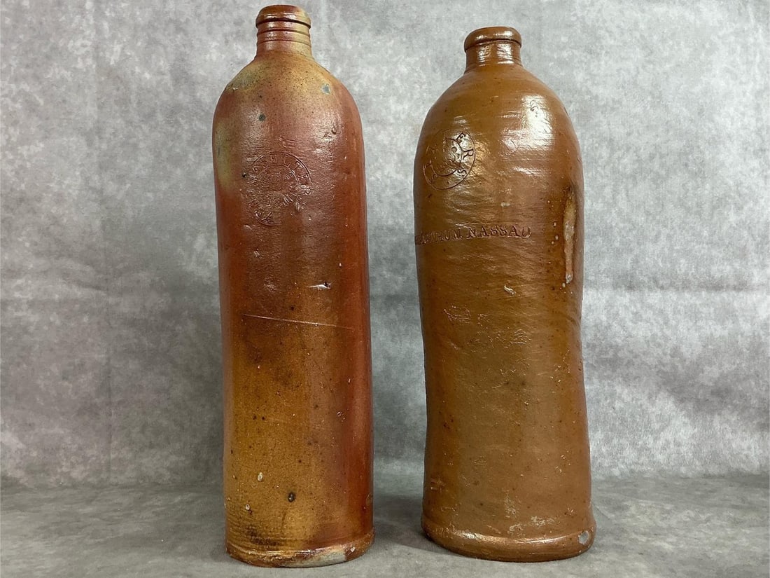 Stoneware Mineral Bottles: Two late 1800's German salt glazed mineral water bottles. Seltzers seal on both, one translates to Duchy of Nassau. Bottles are brown with "thumb" handles. A few spots with chips or wear present. One 