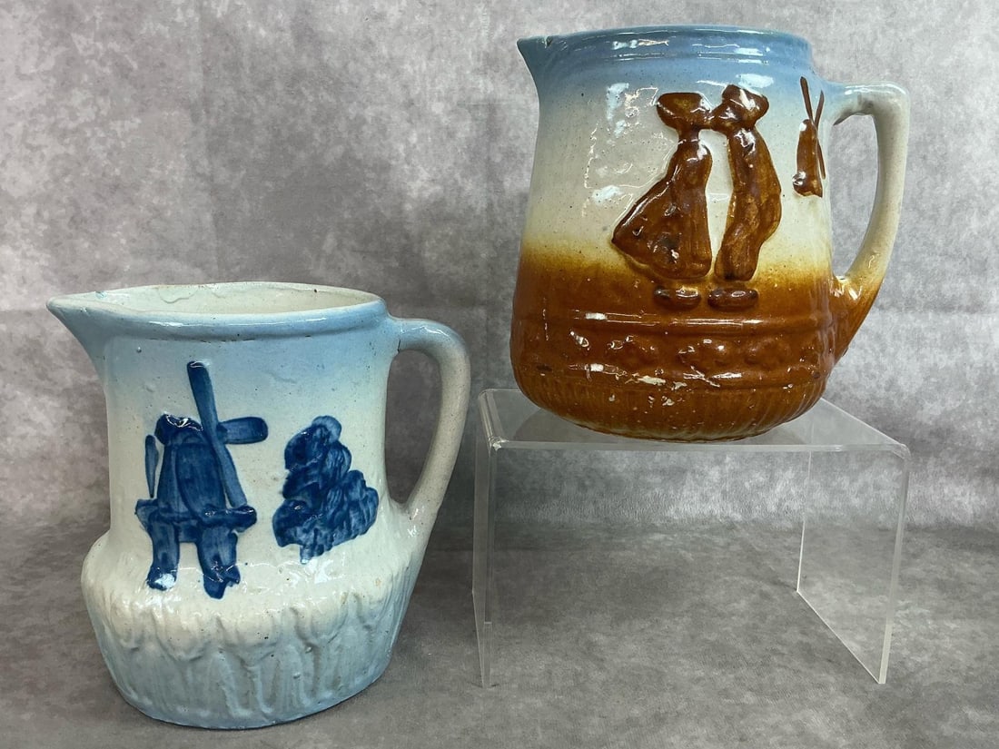 Antique Stoneware Pitchers: Two early 1900's salt glazed stoneware pitchers. Blue and brown Dutch boy and girl, and blue Dutch windmill - both likely Brush-McCoy. Crazing and slight paint loss present. Blue pitcher measures 7