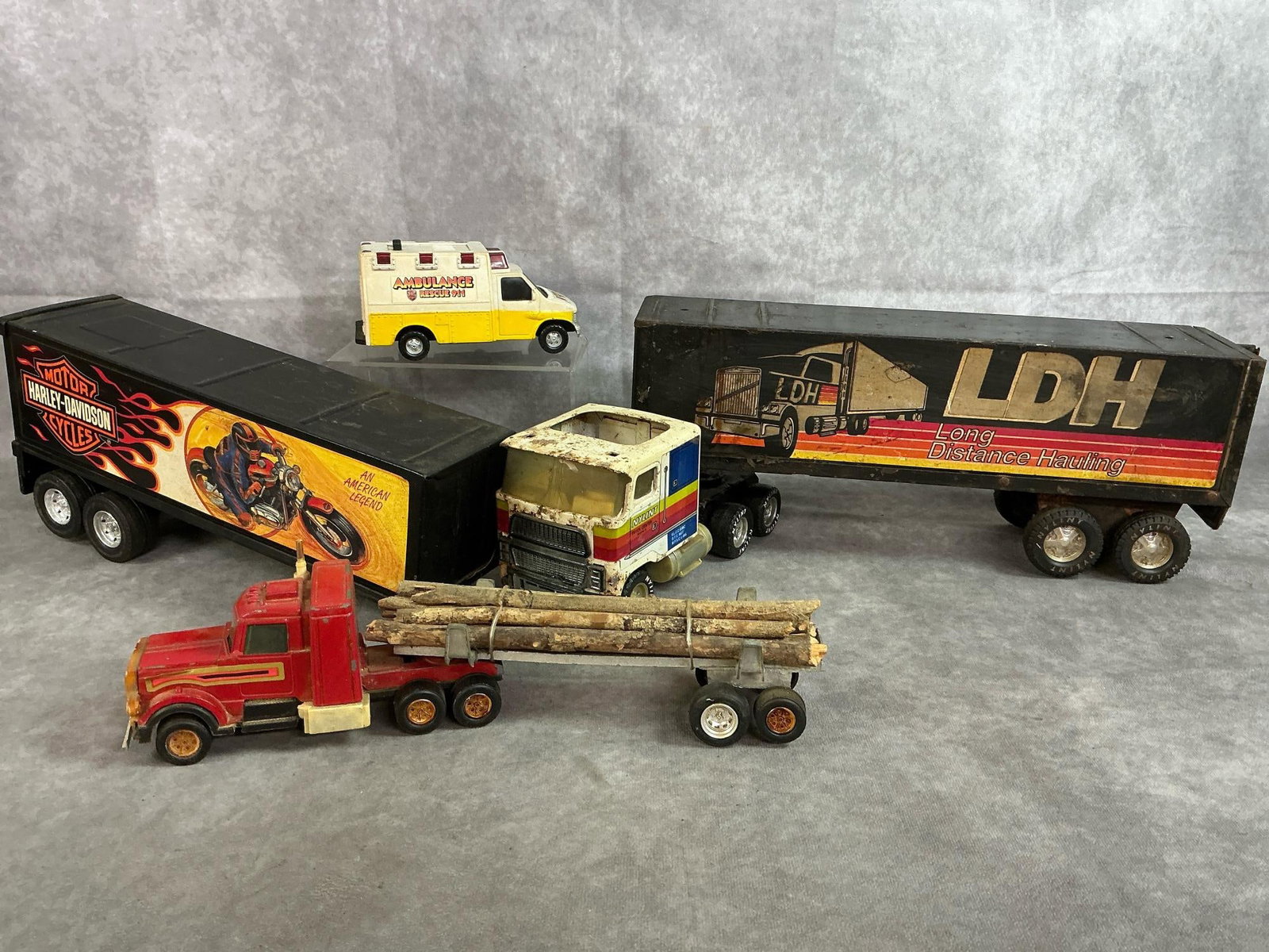 Toy Road Tractors & Ambulance: Four late 1900's toy trucks and trailers. Nylint LDH metalcab and trailer, Harley-Davidson metal trailer, die cast ambulance, and Tootsie Toy log truck. Rust and wear present. LDH truck measures 20.5 