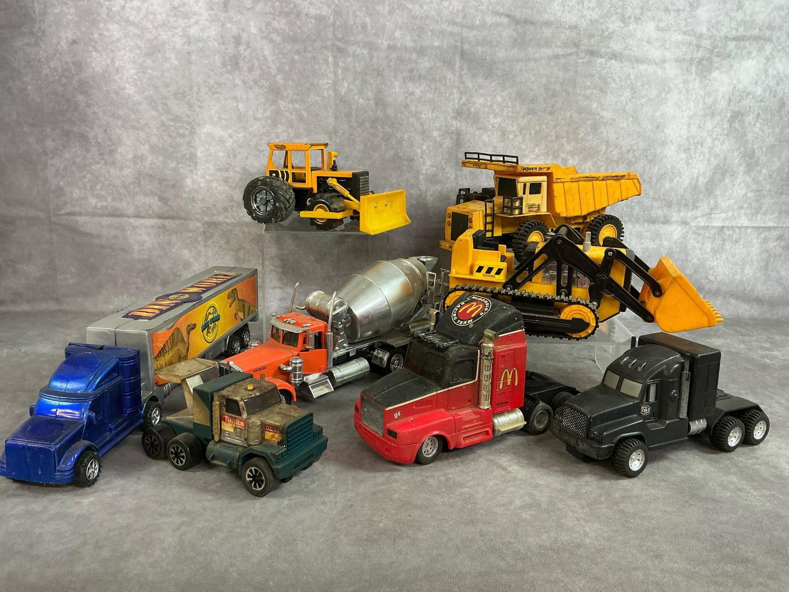 Die-Cast & Plastic Toys: Eight late 1900's toy vehicles. Three plastic construction vehicles, plastic Dino World truck, McDonalds truck and black road tractor. Die cast cement mixer and Tonka truck. Rust and wear present, som