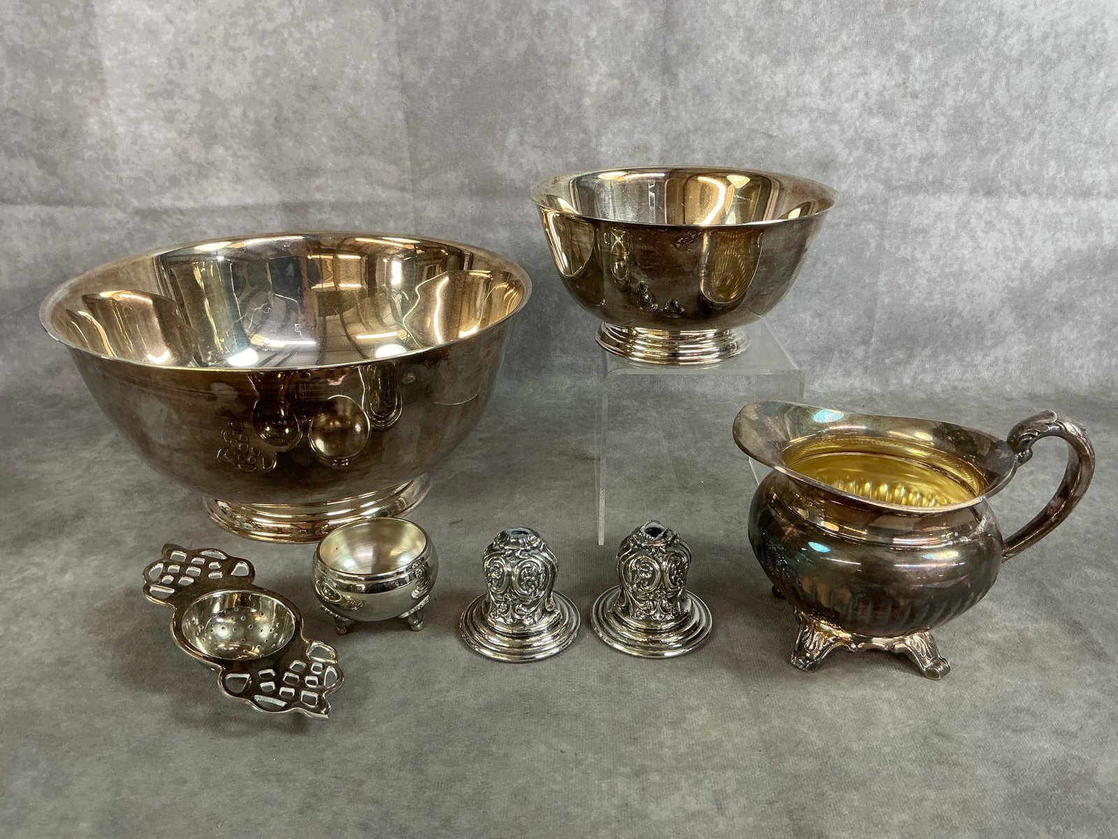 Silver Plated Bowls, Creamer, & More: Seven vintage silver plated serving pieces. Includes two Sons of Liberty reproduction bowls by Oneida Silversmith, tea strainer, miniature candle holders, salt cellar, and creamer. In good condition.