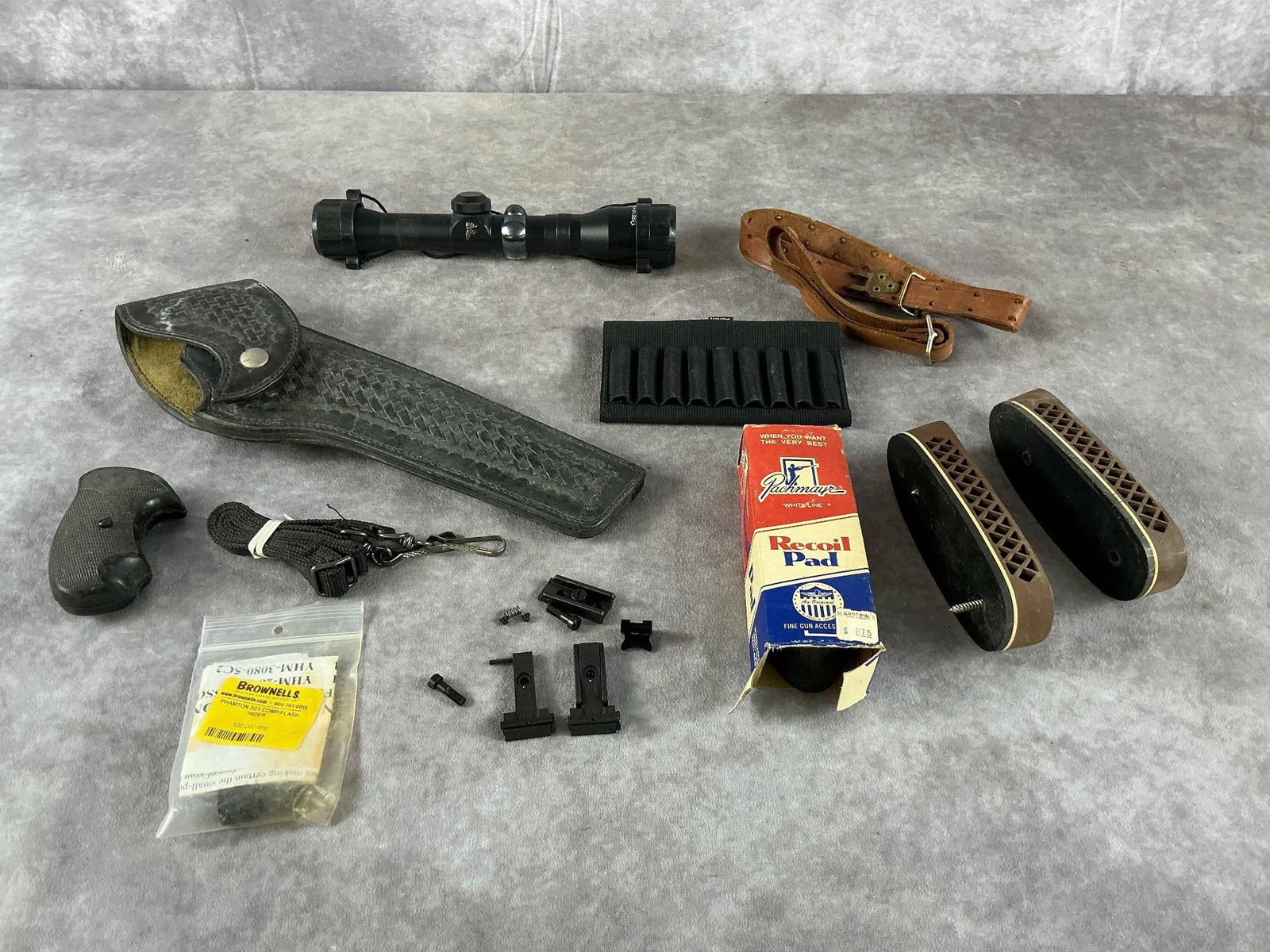 Tasco & Pachmayr Firearm Accessories: Assorted firearm accessories including a Tasco rifle scope, Pachmayr recoil pads, Safariland holster, Allen ammo carrier, and YHM flash hider in mixed black and brown colors. Fair condition with