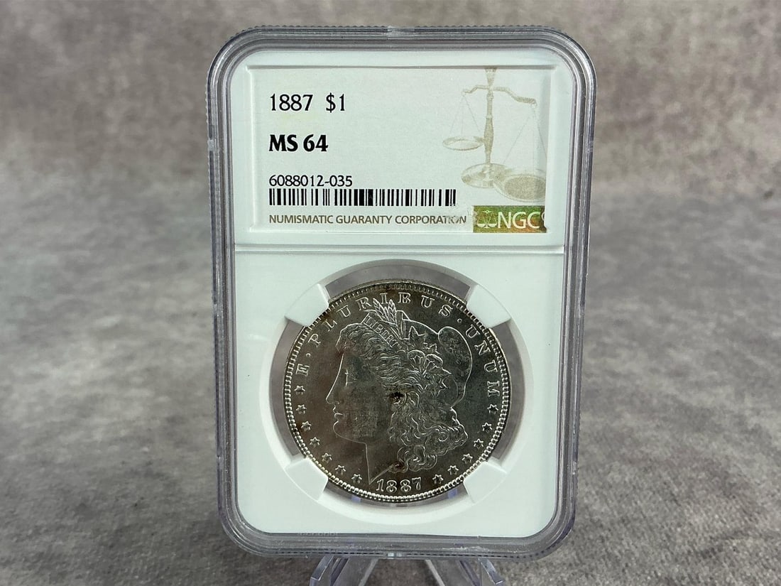 Morgan Dollar: 1887 Morgan Silver Dollar, MS 64, graded by NGC. Certification number 6088012-035. Measures 2.25 inches wide x 3.25 inches tall.