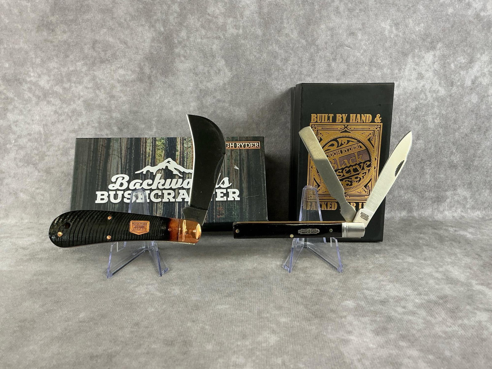 Rough Ryder Knives: 1994 Black Reserve Bearhead Copperhead Rough Ryder pocketknife in original box, and a modern Backwoods Bushcrafter hawkbill Rough Ryder pocketknife in original box. Both in good condition. Largest