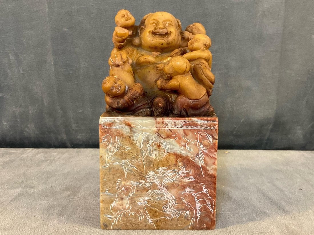 Carved Buddha: Modern Chinese carved soapstone Buddha with playing children over red stone block base with incised landscape and circular seal. Good condition. Measures 3.25 inches wide x 3.25 x 6 inches tall.