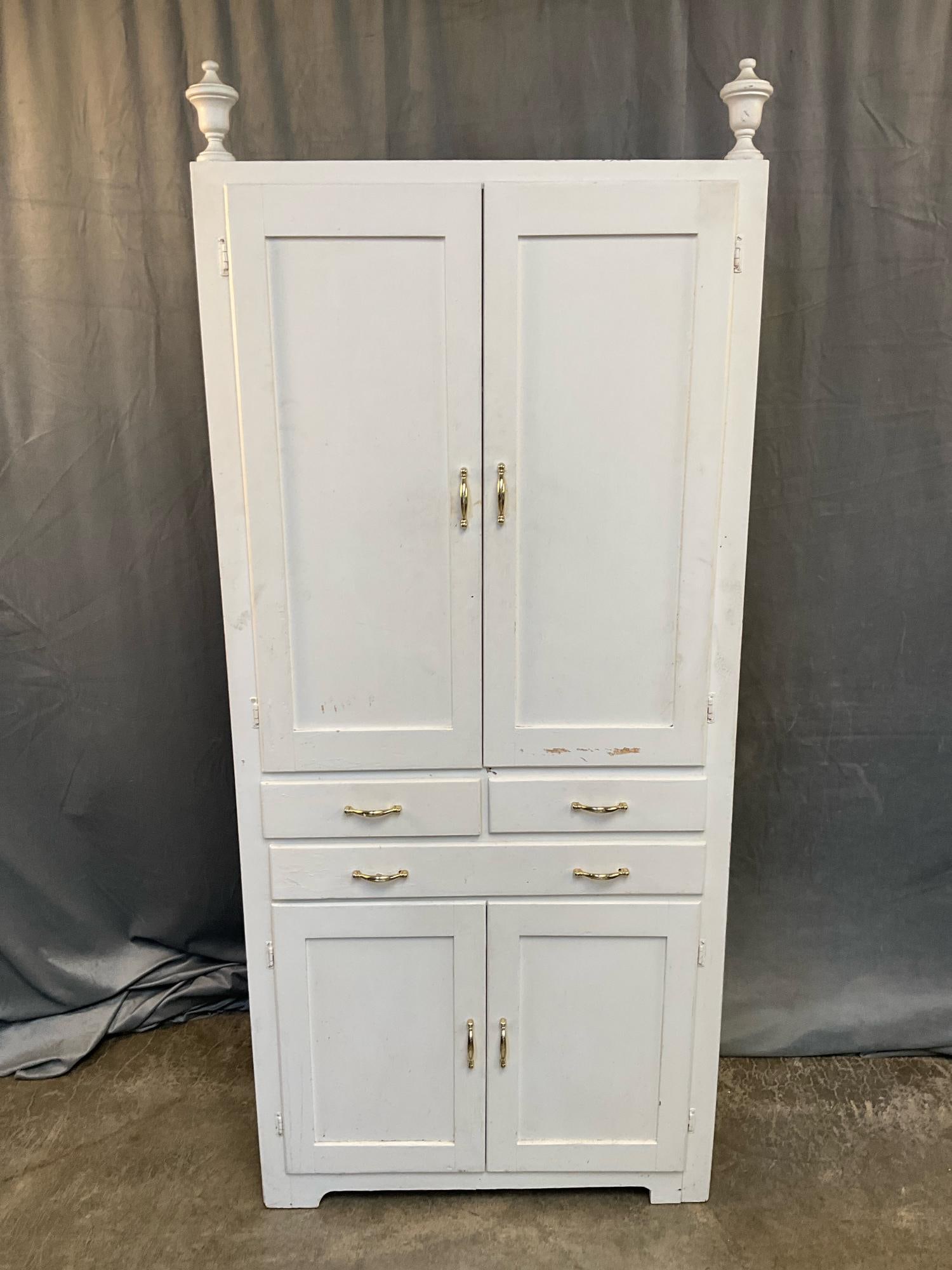 Cupboard: Mid-20th century white painted cupboard with upper and lower storage area, three drawers and urn finials. Repainted, otherwise good condition. Measures 30.25 inches wide x 13.25 x 70 inches tall.