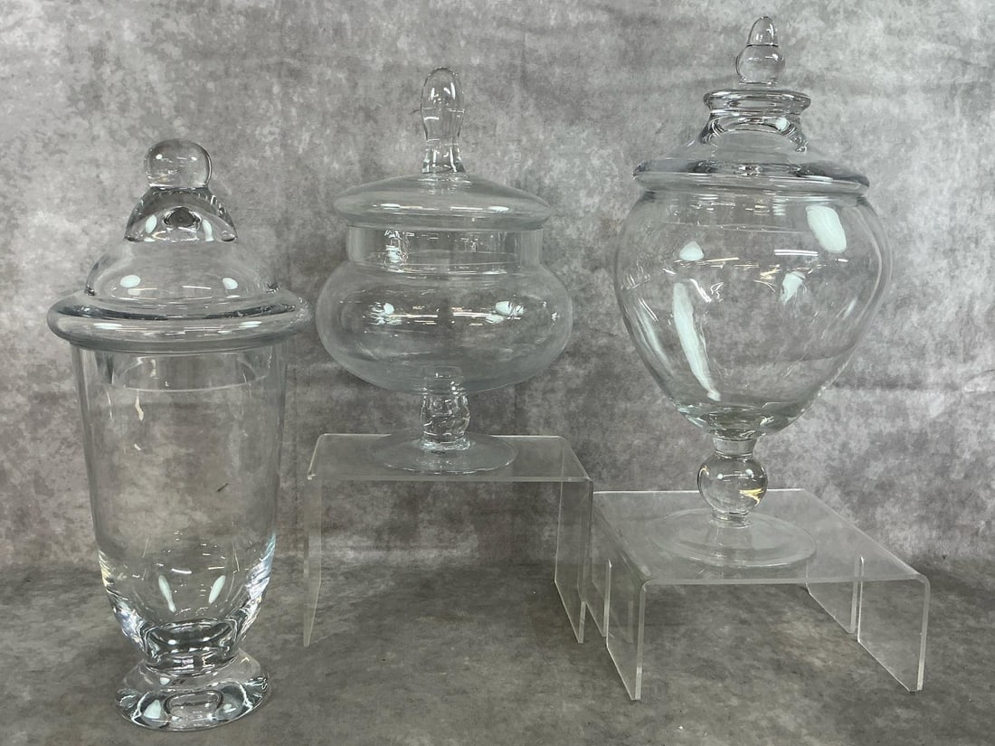 Apothecary Jars: Three late 1900's clear glass apothecary jars with lids. Varied size and shapes on pedestal foot. In good condition. Largest measures 6 inches wide x 12.5 inches tall with lid.