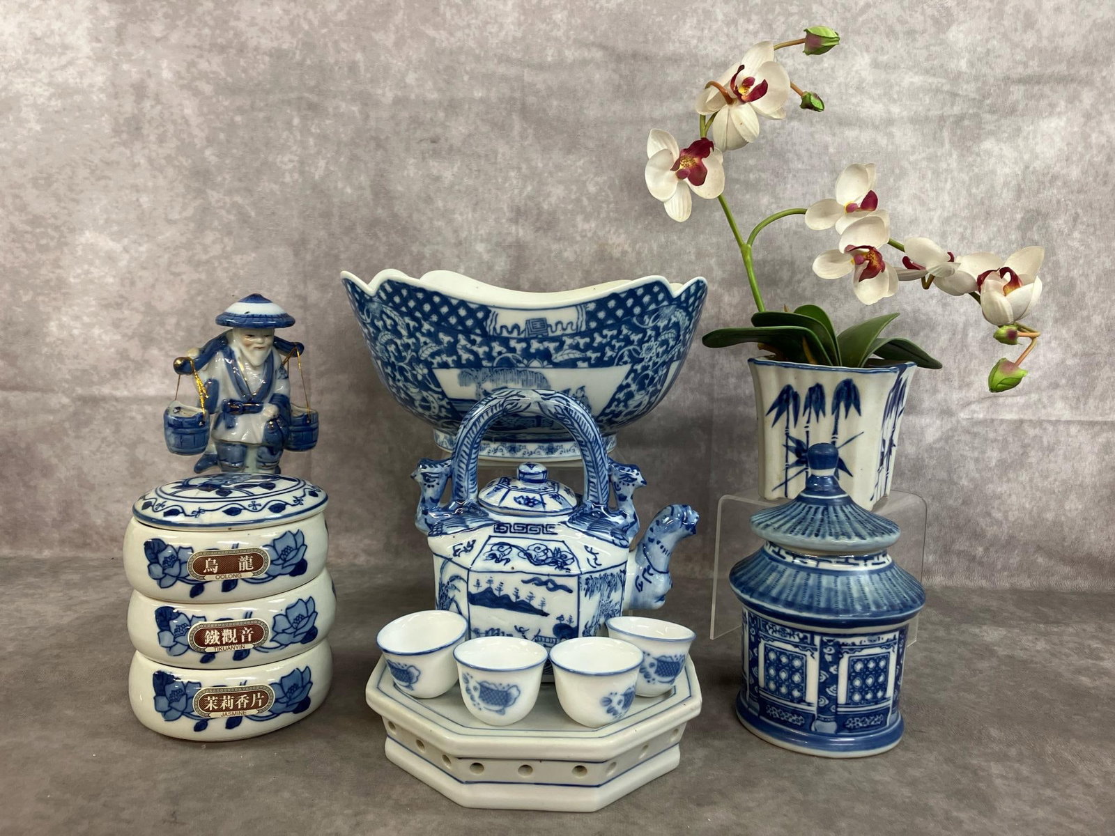 Chinese Blue & White Porcelain: Six vintage blue and white Chinese porcelain pieces. Large bowl with scenes on sides, tea set with tiny cups and base, and porcelain fisherman figure. Includes silk orchids in porcelain pot, and two t
