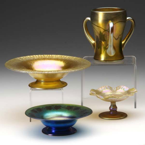 TIFFANY STUDIOS Four Favrile glass pieces: Gold th: TIFFANY STUDIOS Four Favrile glass pieces: Gold three-handled loving cup with green oak leaf and vine decoration, a miniature gold compote, a gold stretched glass footed candy dish, and a blue footed