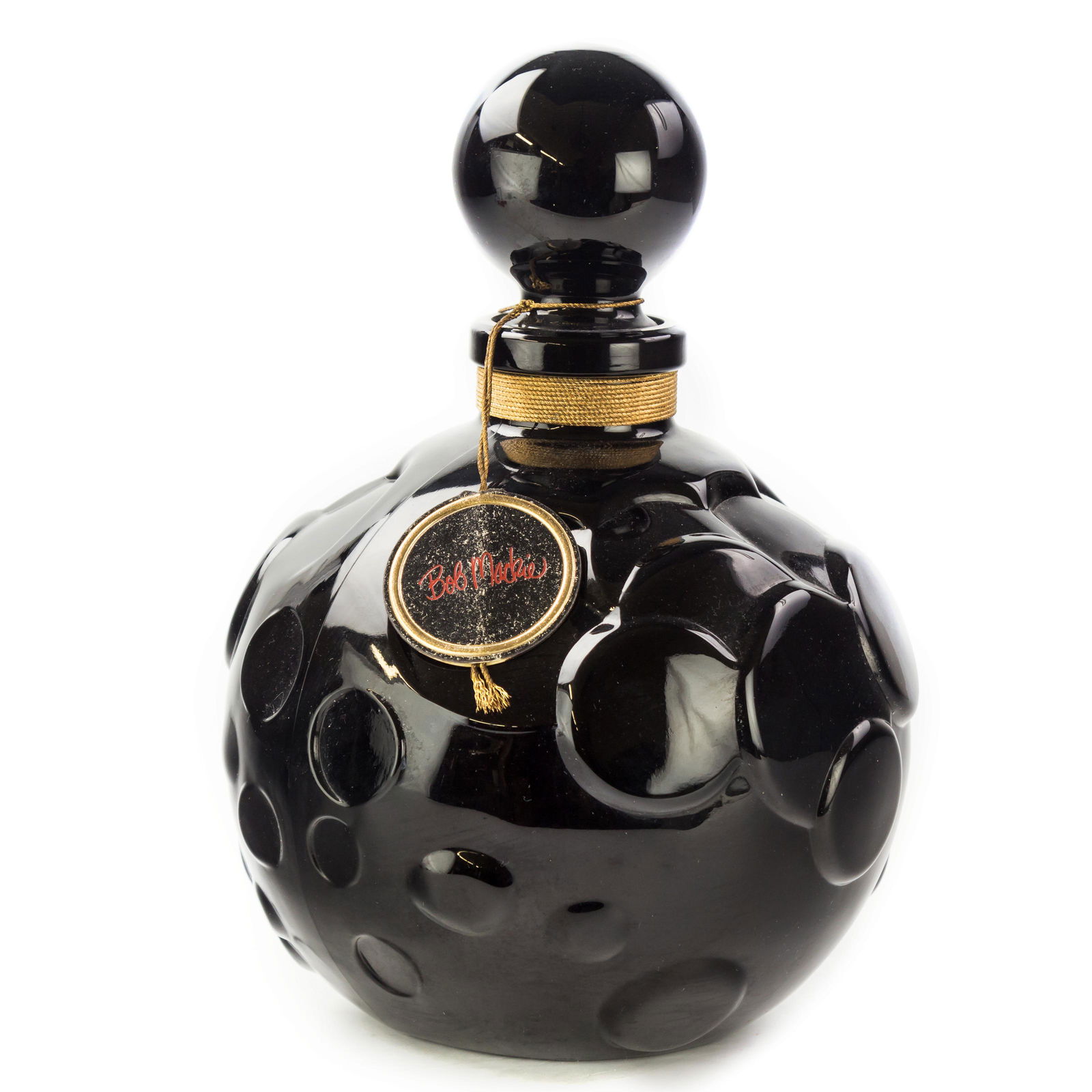 Bob Mackie Oversize Factice Display Perfume Bottle: An oversized counter factice display bottle for Bob Mackie. Black glass with coordinating stopper. Retains original label tag. Measures 11 in. x 8 in. Good condition with mild surface wear. Hill Aucti
