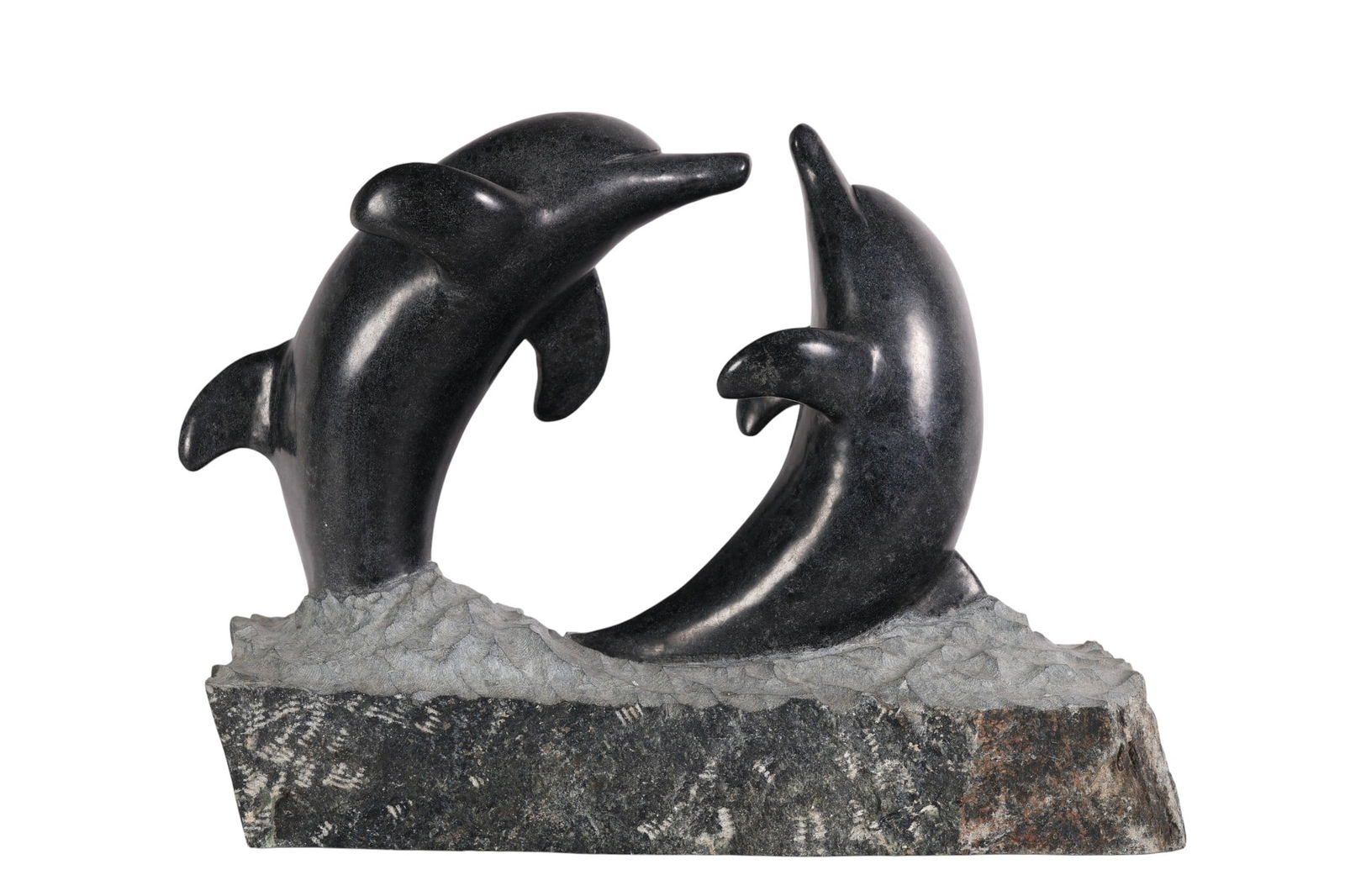 Carved Hardstone Leaping Dolphin Marine Sculpture: Stone sculpture depicting two dolphins in opposing poses, each carved from a dark hardstone with smooth surfaces with mottled designs. Both emerging from a textured base carved to suggest water. No