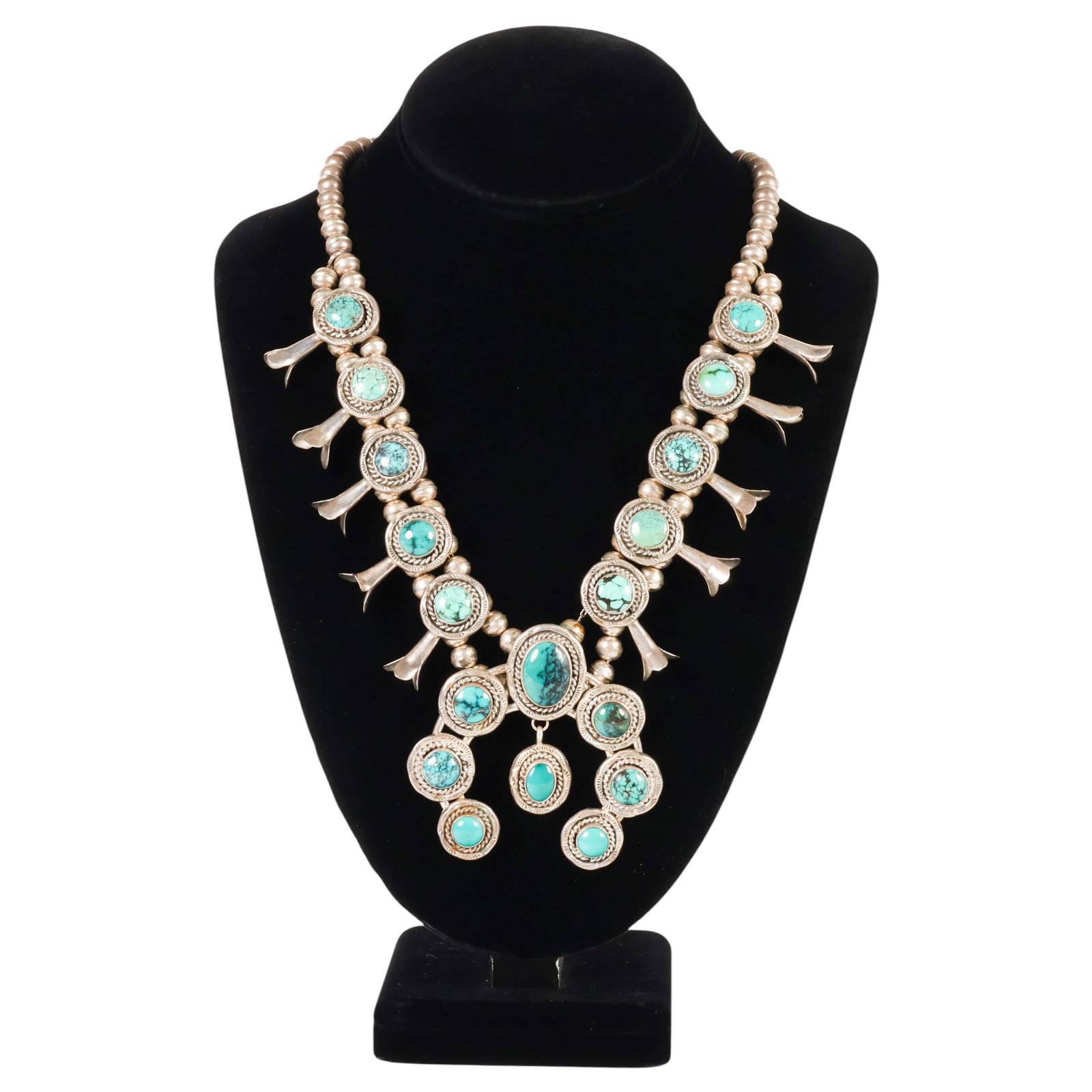 Sterling Silver Turquoise Squash Blossom Necklace: Native American sterling silver squash blossom necklace featuring thirteen bezel-set turquoise cabochons of varying blue-green hues with natural matrix. The necklace is composed of bench-made sterling