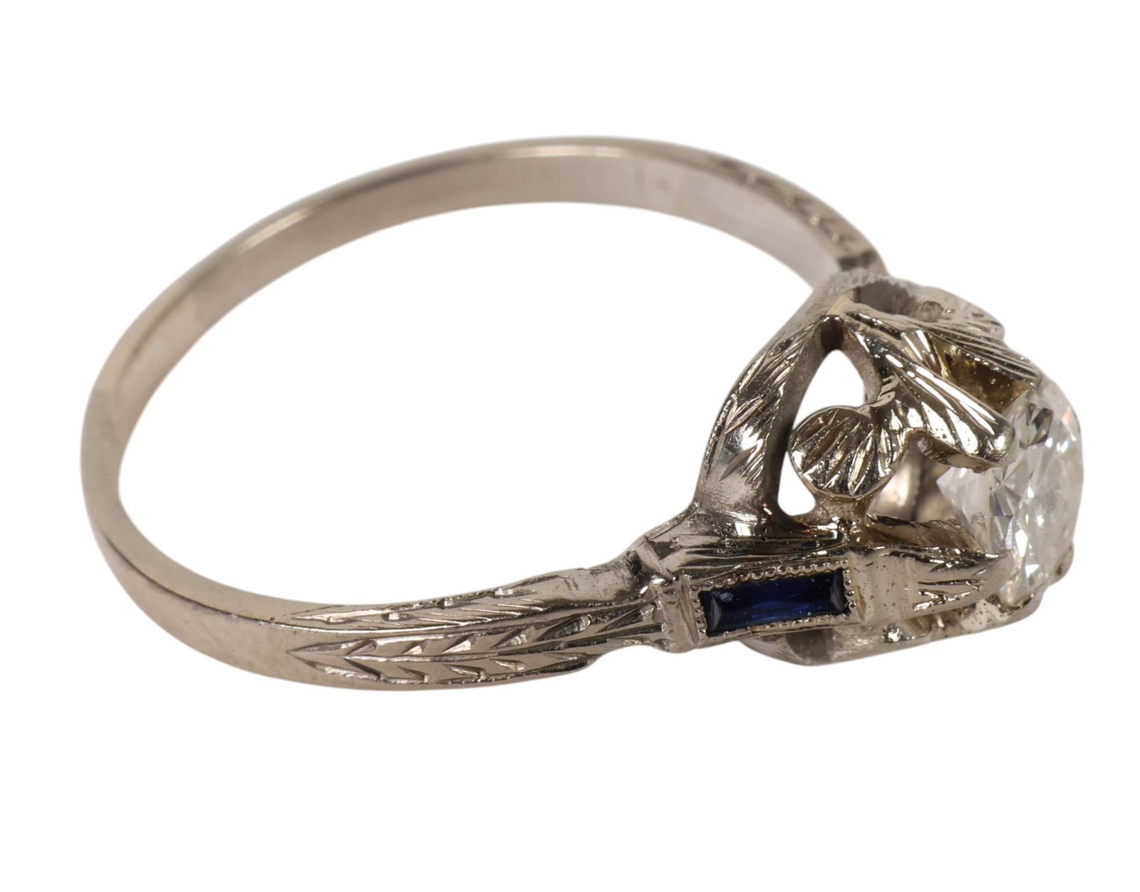 18K Gold Diamond & Sapphire Solitaire Ring Size 8: 18K white gold solitaire ring featuring a round brilliant cut diamond set in a raised prong mount. Engraved shoulders are accented with small channel set dark blue sapphire stones along each side. Ope